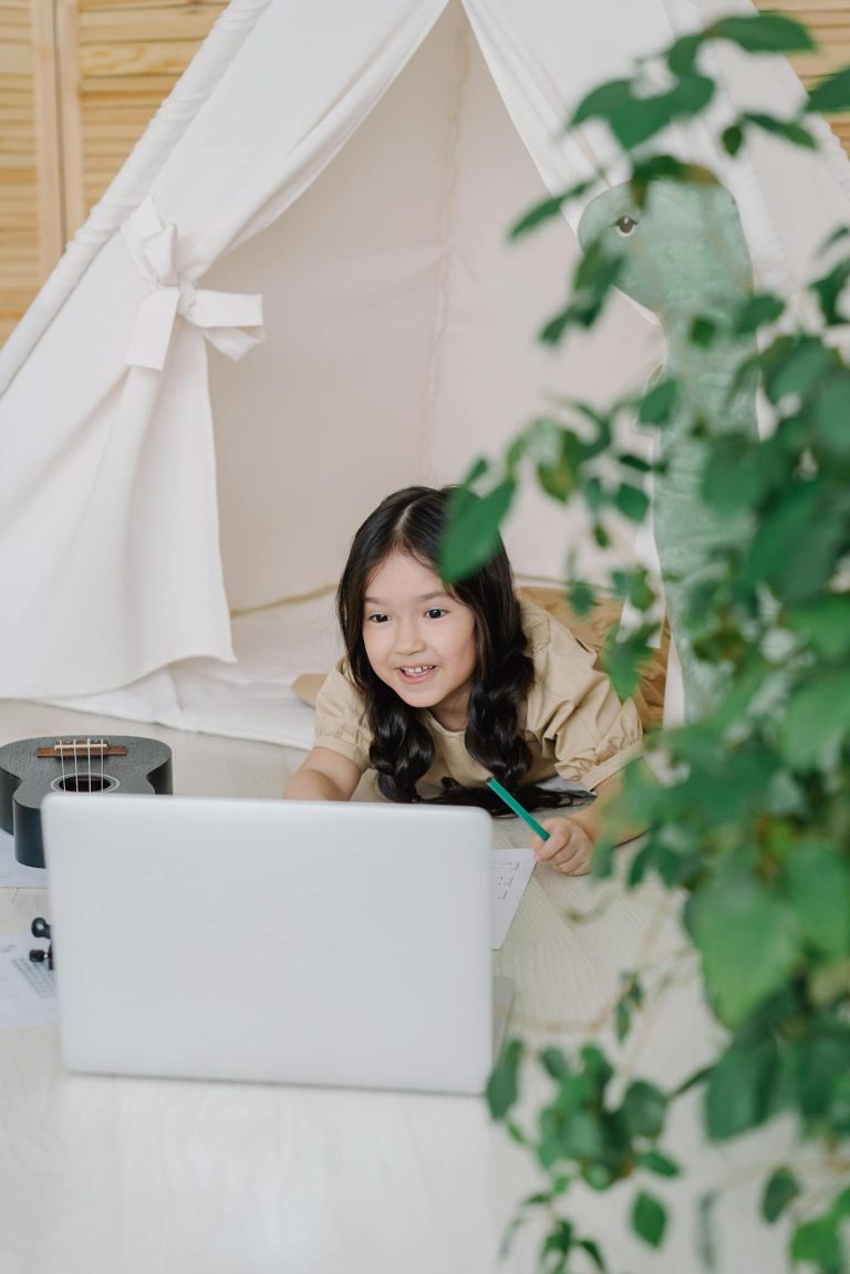 A young girl smiles while interacting with a laptop inside a cozy indoor tent, surrounded by a relaxing home setting.