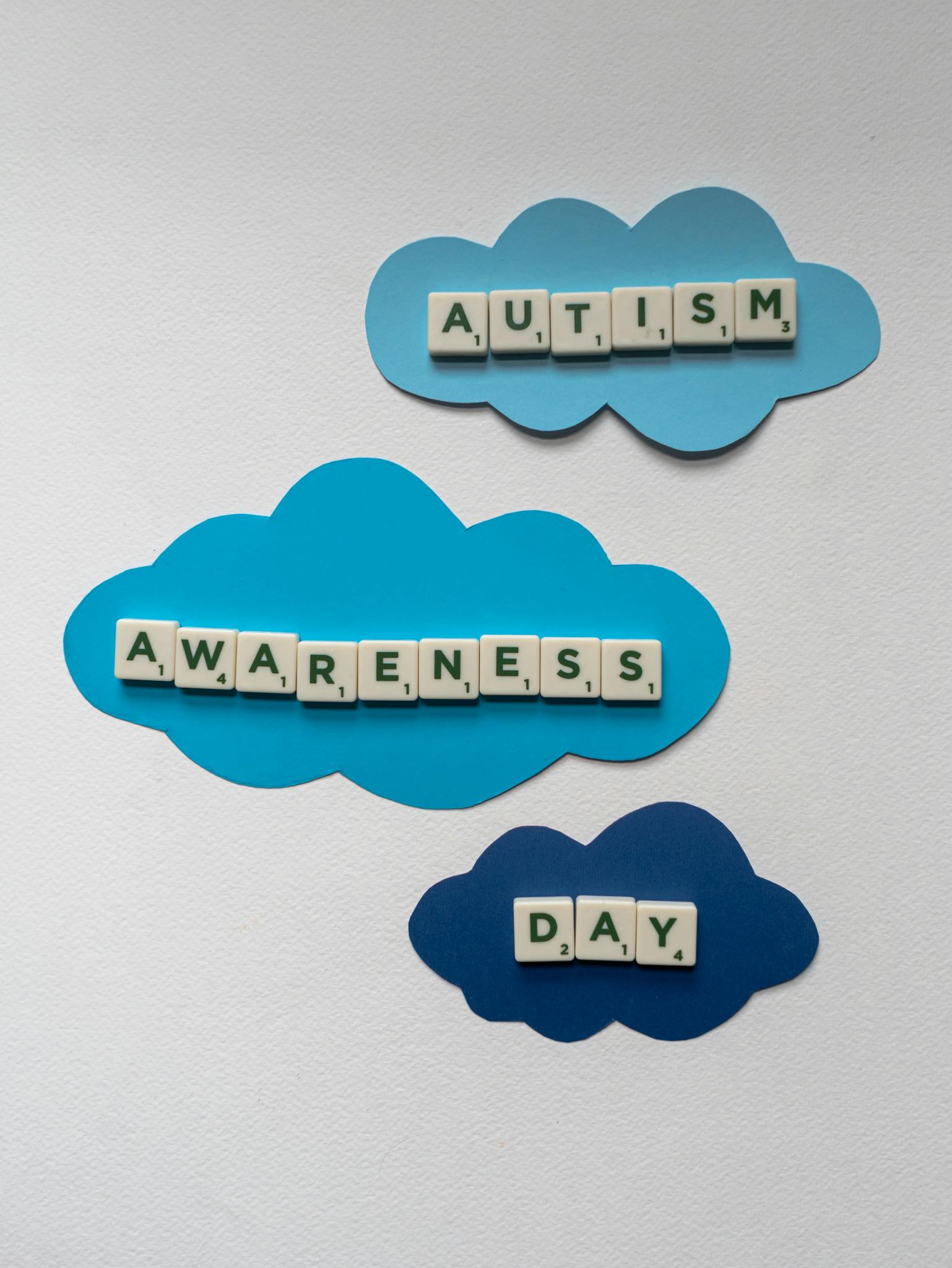Creative display of Scrabble tiles spelling 'Autism Awareness Day' on cloud cutouts.