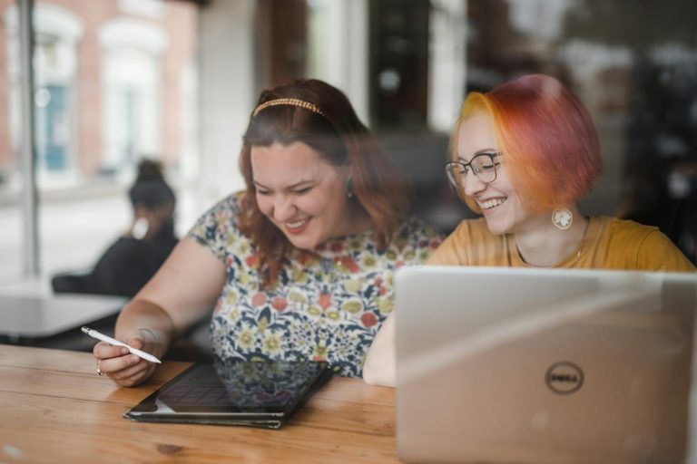 Two women smiling and working together in a cafe, using a laptop and tablet. Collaborative workspace indoors.
